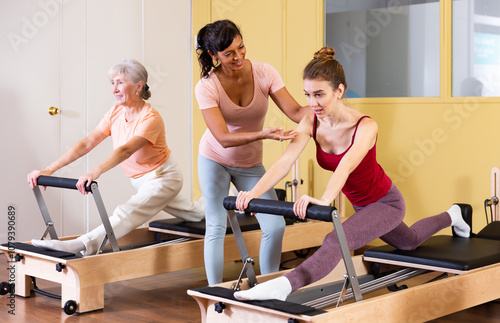 Two health conscious women of different ages perform a Pilates exercise using a reformer bed, where a female instructor ..helps them do it correctly photo