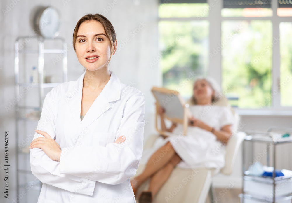Fototapeta premium Smiling young lady wearing medical scrub standing at treatment room in front of female patient lying at examination couch.