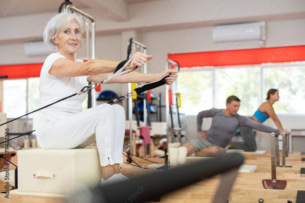 Obraz premium Senior woman works out on in gym on Pilates simulator. Sports club client on reformer with cable and rope performs exercise to stretch muscles of arms. Movement in all directions