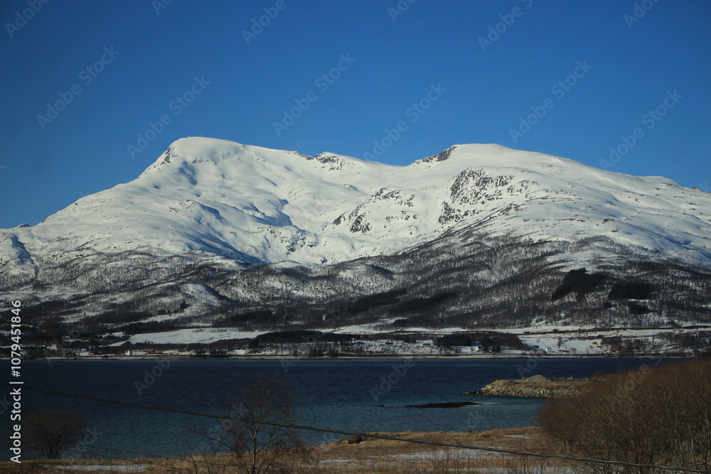 Naklejka premium Winter landscapes in Norway showcase serene snowy mountains, icy fjords, and peaceful coastal views. Capturing the beauty of snow-covered trees, quiet roads, and glowing sunsets creates a sense of cal