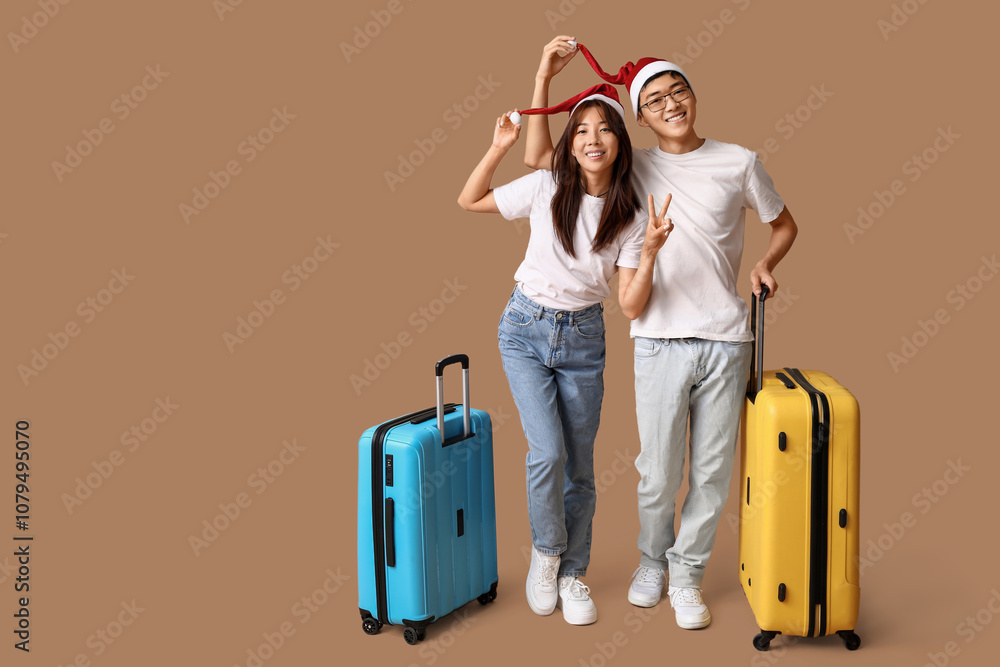 Beautiful young Asian couple in Santa hats with suitcases on brown background. Christmas celebration Beautiful young Asian couple in Santa hats with suitcases on brown background. Christmas celebration