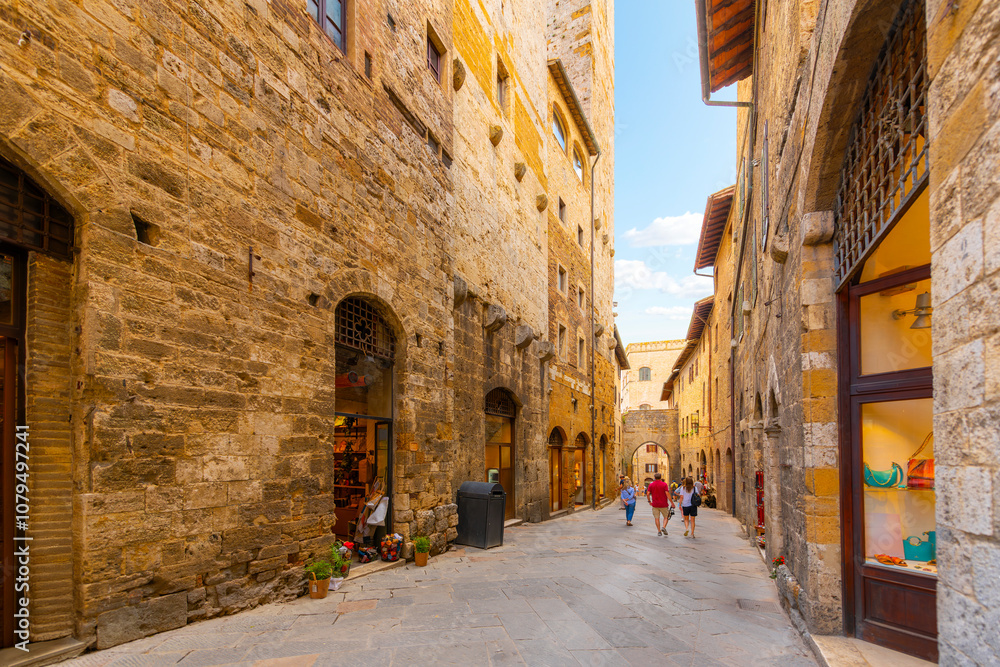 Fototapeta premium Tourists walk through a narrow stone street of cafes and shops in the historic center of the Tuscan Hill Town of San Gimignano, Italy.
