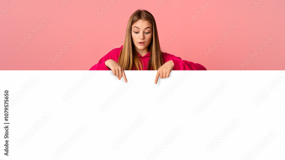 Beautiful teenager girl looks and shows to fingers down on pink background, studio shot, free space Beautiful teenager girl looks and shows to fingers down on pink background, studio shot, free space