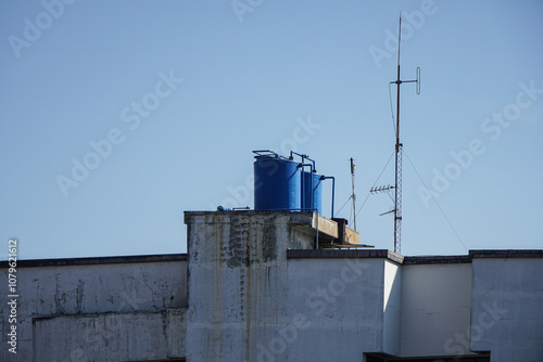 Three blue water tanks on a rooftop, with a tall antenna. The building is made of concrete and has a white paint job. Blue Water Tanks on a Rooftop Background. photo