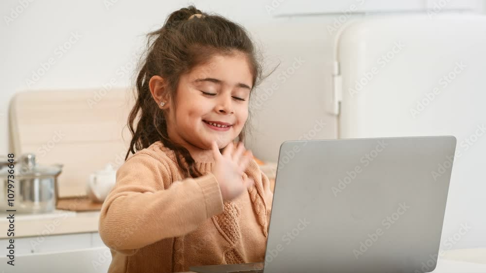 A cheerful girl with long hair waves at her laptop screen while sitting in a bright kitchen. Her smile shows joy, indicating she is engaging in a virtual conversation. A cheerful girl with long hair waves at her laptop screen while sitting in a bright kitchen. Her smile shows joy, indicating she is engaging in a virtual conversation.