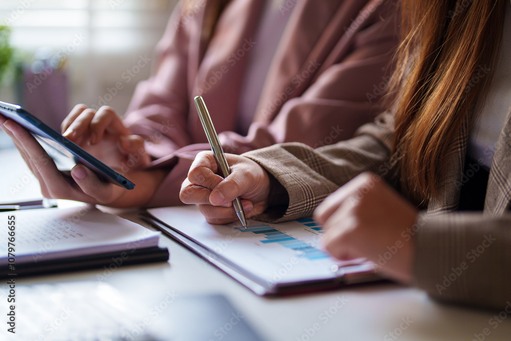 Two businesswomen sit together in an office setting, with one showing something on her smartphone to the other. They are engaged and focused. Two businesswomen sit together in an office setting, with one showing something on her smartphone to the other. They are engaged and focused.