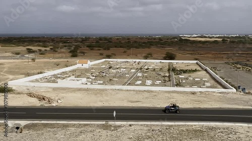 Elevated perspective of Rabil Cemetery on Boa Vista, Cape Verde, set amidst a barren desert with distant ocean views, capturing the stark contrast of the white walls against the rugged surroundings photo