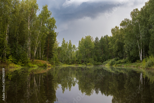 Summer landscape with a forest lake. Evening view of a small lake in the forest. Birches grow along the banks. Trees are reflected in the water. Cloudy weather. Clouds in the sky. Outdoor recreation.