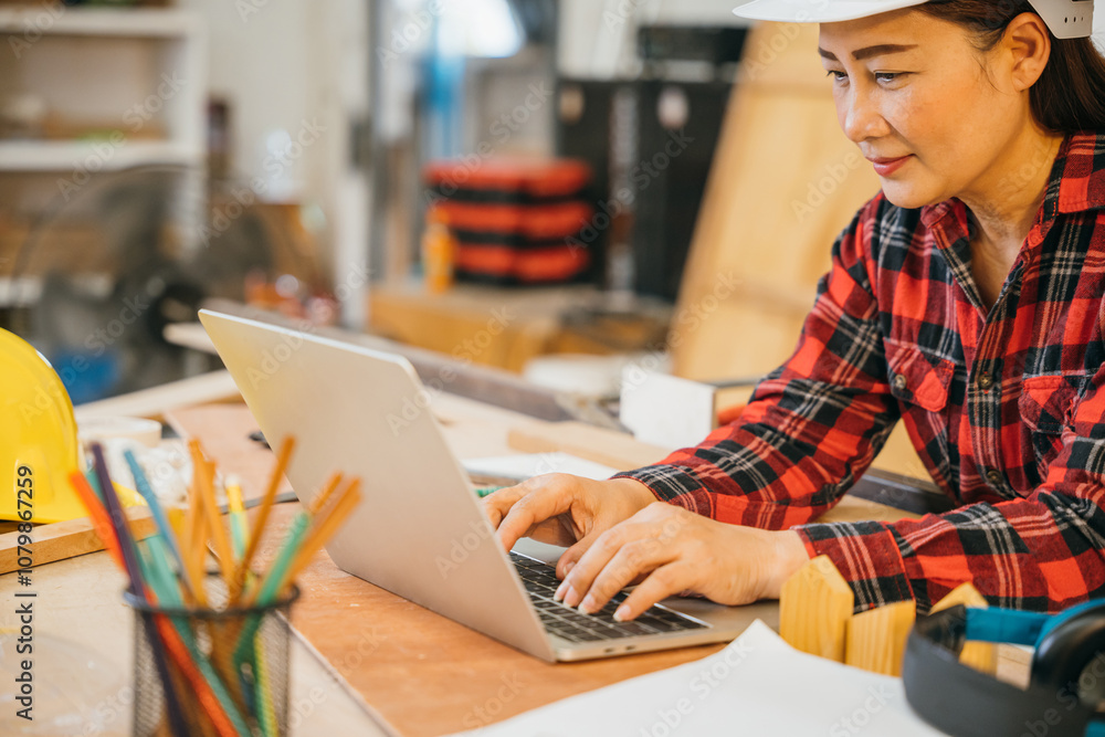 Carpenter woman in a white hardhat and red plaid shirt uses a laptop in a woodshop, surrounded by tools, wood scraps, and pencils. Ideal for online learning, client orders, or woodworking projects Carpenter woman in a white hardhat and red plaid shirt uses a laptop in a woodshop, surrounded by tools, wood scraps, and pencils. Ideal for online learning, client orders, or woodworking projects