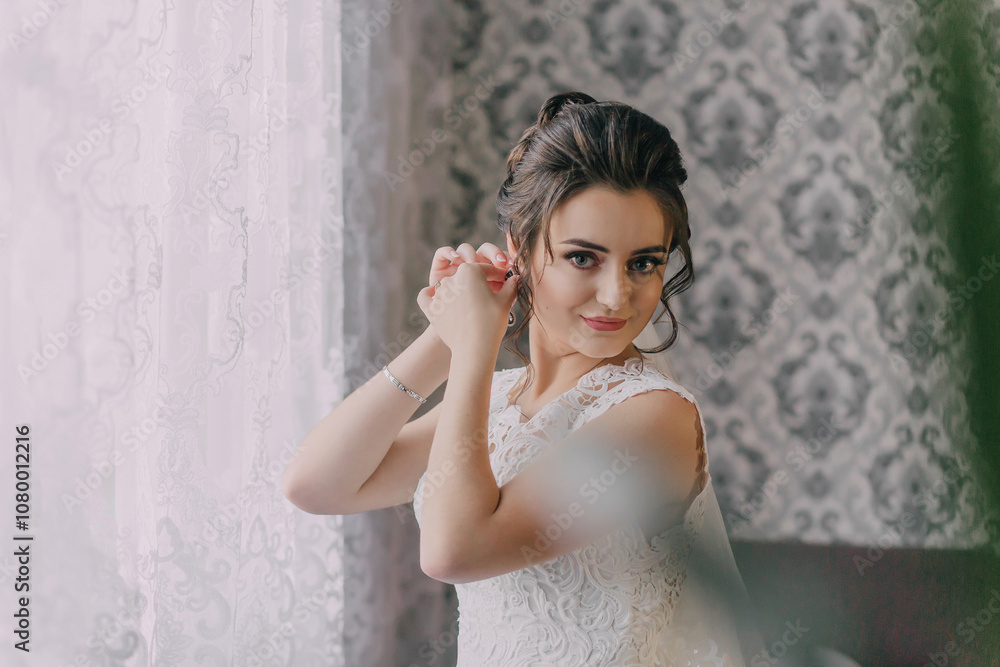 Fototapeta premium A woman is getting ready for a wedding, putting on her wedding dress and earrings. She is standing in front of a window, and the room has a vintage feel to it