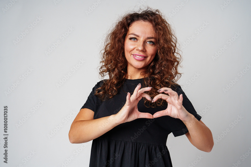 A young woman with curly hair makes a heart shape with her hands against a neutral backdrop A young woman with curly hair makes a heart shape with her hands against a neutral backdrop