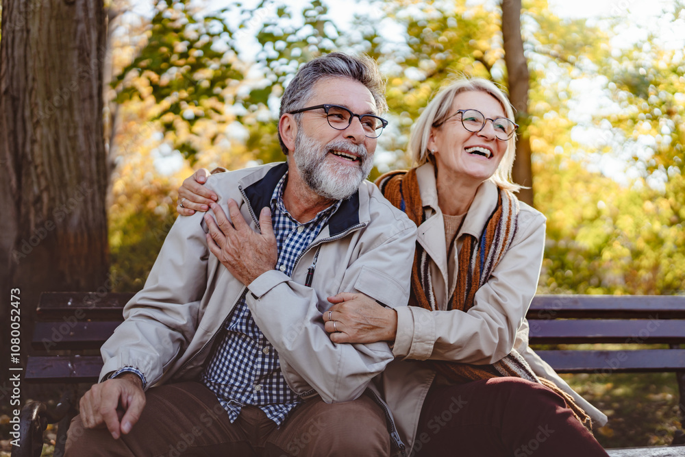 Fototapeta premium Happy senior couple sitting on a park bench in autumn, smiling and enjoying a sunny day outdoors with colorful leaves in the background.