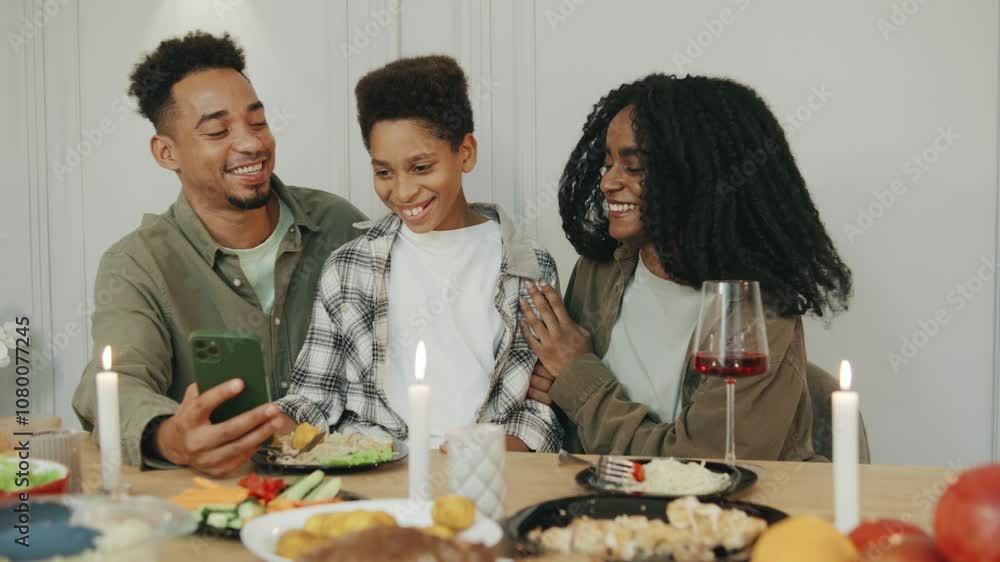 Around the dinner table, an African American family shares laughter while watching something on a smartphone, celebrating joyful holiday moments together. Around the dinner table, an African American family shares laughter while watching something on a smartphone, celebrating joyful holiday moments together.