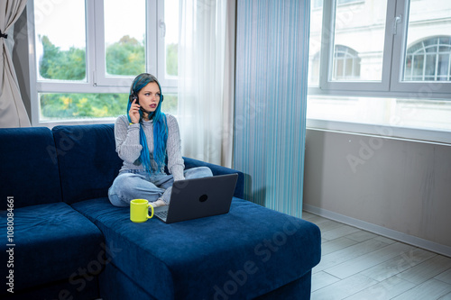 Beautiful blue haired hipster woman is working from home sitting on her couch, wearing headphones with microphone to attend a meeting with laptop. Concept of working from home, using technology. photo