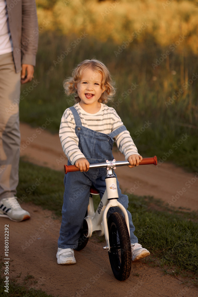 Young father teaching happy beautiful little girl to ride on first bike without pedals on sidewalk at city park. Learning to keep balance. Cute 3 years old toddler. Warm summer day. Front view. Young father teaching happy beautiful little girl to ride on first bike without pedals on sidewalk at city park. Learning to keep balance. Cute 3 years old toddler. Warm summer day. Front view.