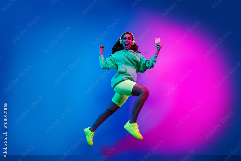 Fototapeta premium Full length photo of charming positive woman wear sweatshirt headphones jumping winning game device isolated neon blue color background