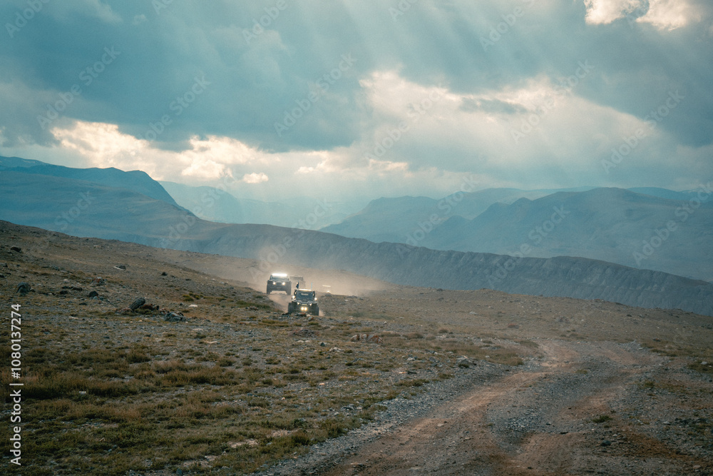 Obraz premium Buggy trip in a Altai desert. Mountain landscape with hills and beautiful blue sky with clouds in background. Off-road vehicles driving on a dust dirt road. Active leisure in Altai Republic.