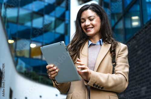 Successful businesswoman using a digital tablet while standing in front of business office building. Young businessperson, professional occupation worker, modern technology developer on urban street.