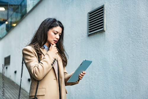 Successful businesswoman using a digital tablet while standing in front of business office building. Young businessperson, professional occupation worker, modern technology developer on urban street.