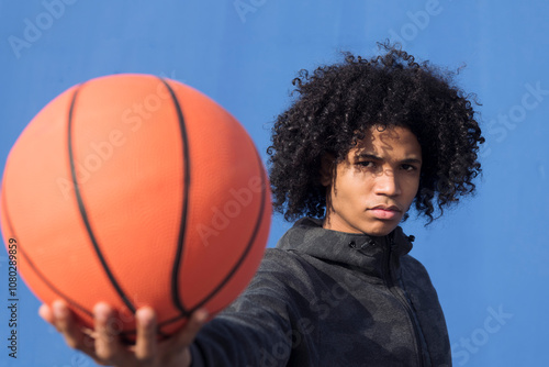 serious unemotional teen african american male with curly hair offering hand with basketball ball towards camera against blue background, concept of sport and young lifestyle, copy space for text photo