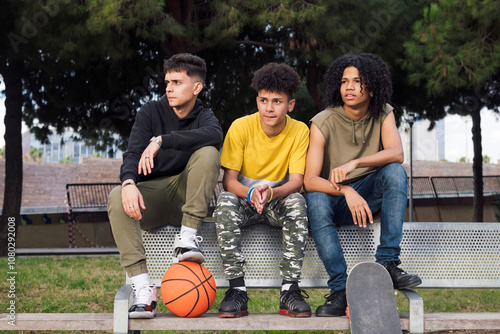 multiracial teen male friends sitting on bench of a park with basketball and skateboard, concept of youth and urban lifestyle