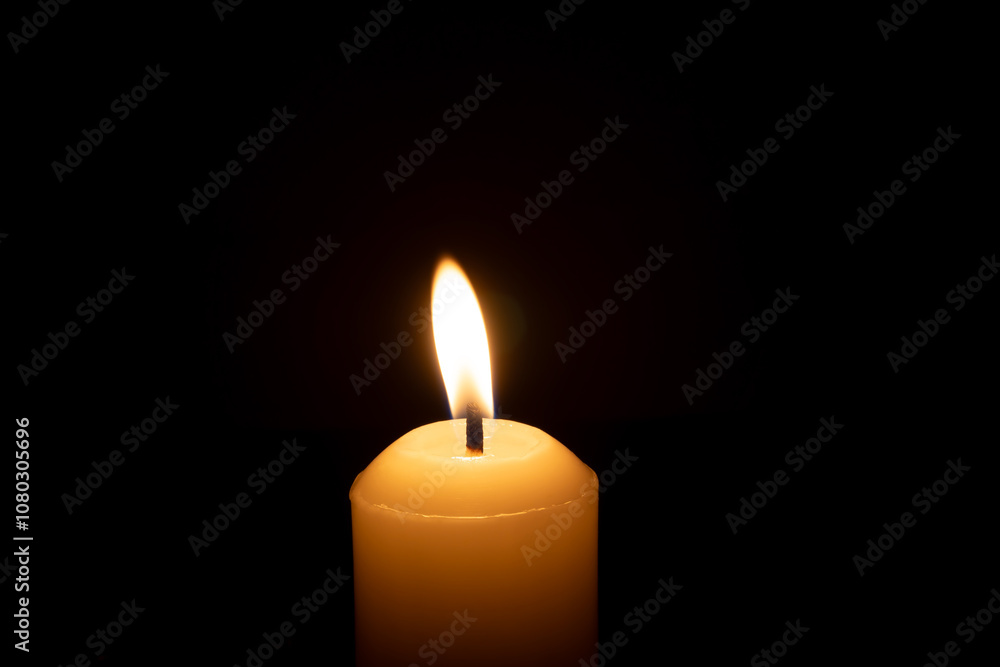 A single burning candle flame or light glowing on a big white candle isolated on black or dark background on table in church for Christmas, funeral or memorial service A single burning candle flame or light glowing on a big white candle isolated on black or dark background on table in church for Christmas, funeral or memorial service