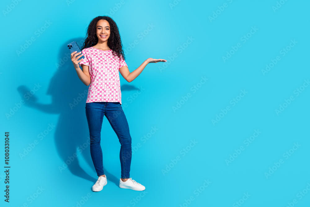 Full length photo of cheerful pretty lady dressed pink t-shirt chatting modern device showing arm empty space isolated blue color background Full length photo of cheerful pretty lady dressed pink t-shirt chatting modern device showing arm empty space isolated blue color background