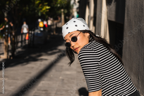 Stylish young asian woman with bandana and sunglasses poses against a wall in a city street