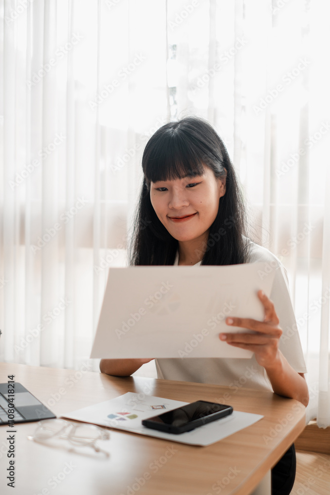A young woman sits at a desk reviewing documents, surrounded by modern technology and natural light from a window. A young woman sits at a desk reviewing documents, surrounded by modern technology and natural light from a window.