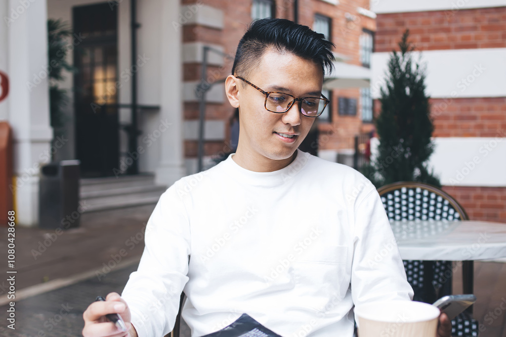 Fototapeta premium Adult ethnic man using smartphone while sitting at table with notepad and to go beverage on cafe terrace