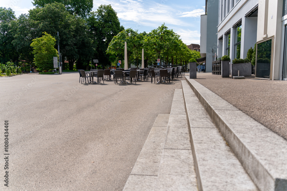 Naklejka premium Place in the city with empty chairs and tables at the outside in front of a cafe. Scene viewed from some distance. Beautiful day with blue, slightly clouded sky.