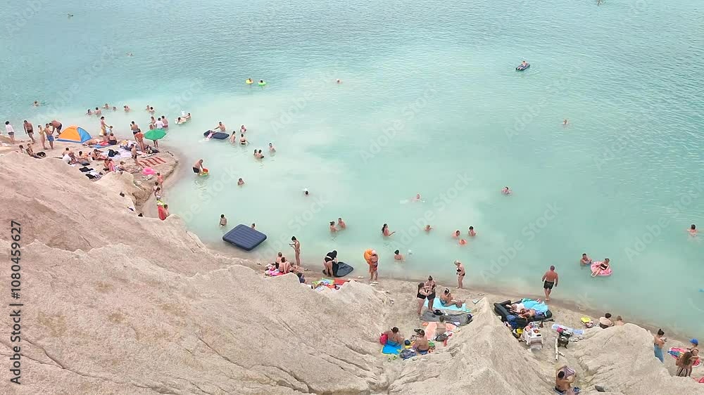 Many people swimming in the blue water near the sandy shore in a flooded quarry on a summer day. Beautiful blue water of large lake and many people swimming near the sandy shore of the slope. Top view Many people swimming in the blue water near the sandy shore in a flooded quarry on a summer day. Beautiful blue water of large lake and many people swimming near the sandy shore of the slope. Top view