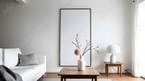 A minimalist living room with a large blank vertical frame hanging on a white wall, positioned above a low wooden coffee table with a white vase holding a few dried branches and a pinkish-white flower