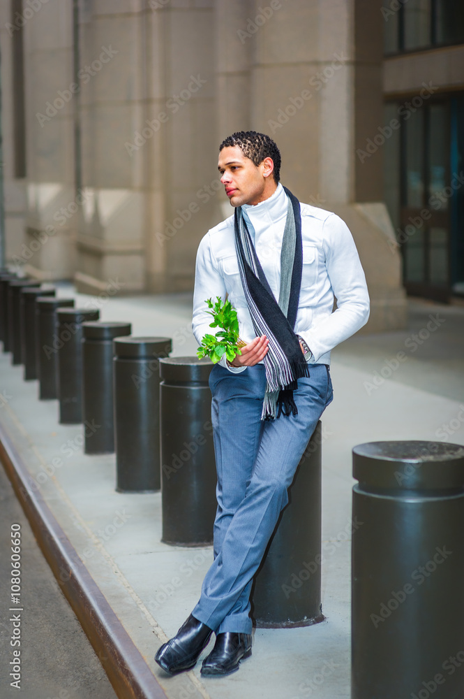 A person leans against a series of cylindrical posts on a city street, holding a sprig of greenery. Dressed in a white shirt, grey trousers, and a black scarf, they appear thoughtful.