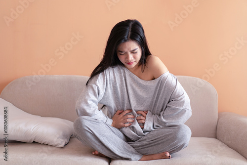 Wall Mural Asian girl sitting on the sofa holding her stomach in discomfort