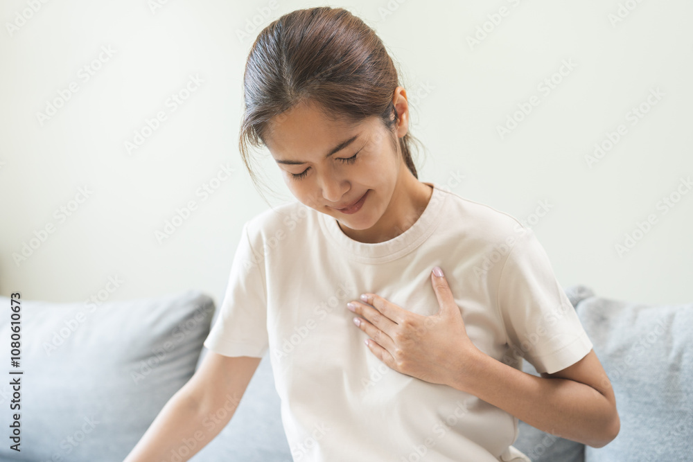Heart attack, health care concept, suffering acute cardiac asian young woman hand touching on her chest pain, breathing problem, asthma attack, pressure while sitting on sofa with closed eyes at home. Heart attack, health care concept, suffering acute cardiac asian young woman hand touching on her chest pain, breathing problem, asthma attack, pressure while sitting on sofa with closed eyes at home.