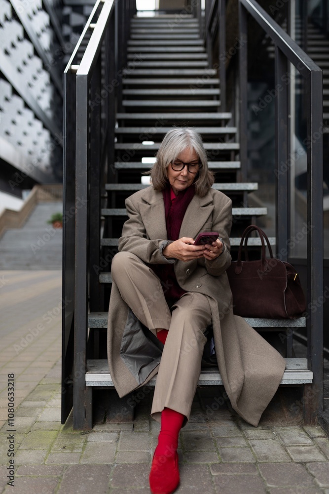 Middle-aged businesswoman in a tailored beige suit sits on stairs checking her phone in an urban setting during the day Middle-aged businesswoman in a tailored beige suit sits on stairs checking her phone in an urban setting during the day