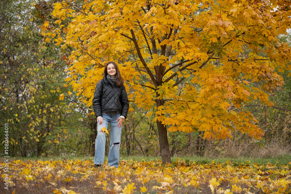 A young woman stands happily near a tree adorned with bright yellow leaves, surrounded by a carpet of fallen foliage in a tranquil autumn park setting. A young woman stands happily near a tree adorned with bright yellow leaves, surrounded by a carpet of fallen foliage in a tranquil autumn park setting.
