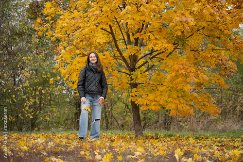 A young woman stands happily near a tree adorned with bright yellow leaves, surrounded by a carpet of fallen foliage in a tranquil autumn park setting.