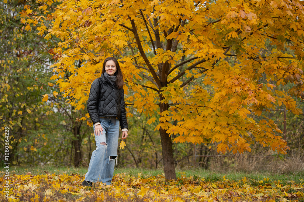 A woman stands beside a striking golden maple tree in a park, dressed warmly as autumn leaves cover the ground, capturing the beauty of the season. A woman stands beside a striking golden maple tree in a park, dressed warmly as autumn leaves cover the ground, capturing the beauty of the season.