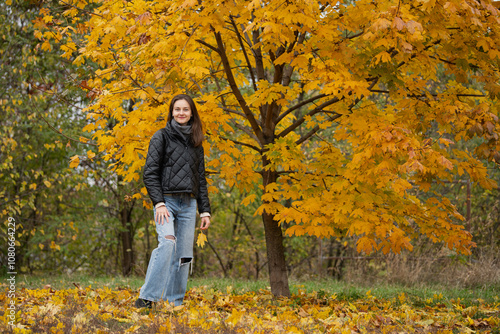 A woman stands beside a striking golden maple tree in a park, dressed warmly as autumn leaves cover the ground, capturing the beauty of the season.