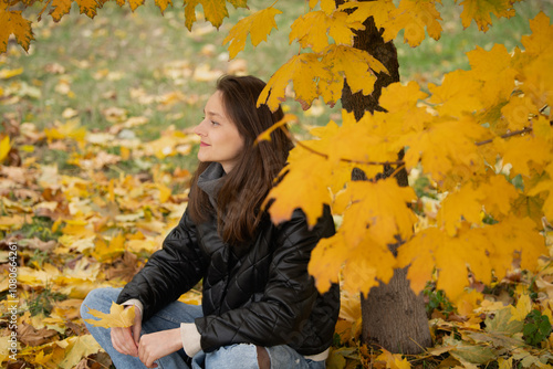 A young woman enjoys a peaceful moment sitting beneath a tree with bright yellow leaves, surrounded by colorful autumn foliage in a tranquil park.
