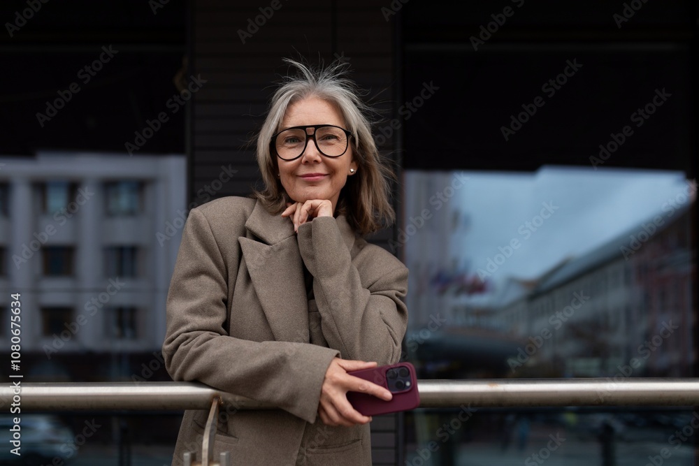 Fototapeta premium Senior businesswoman in casual attire stands confidently outside an urban building while holding her phone in hand