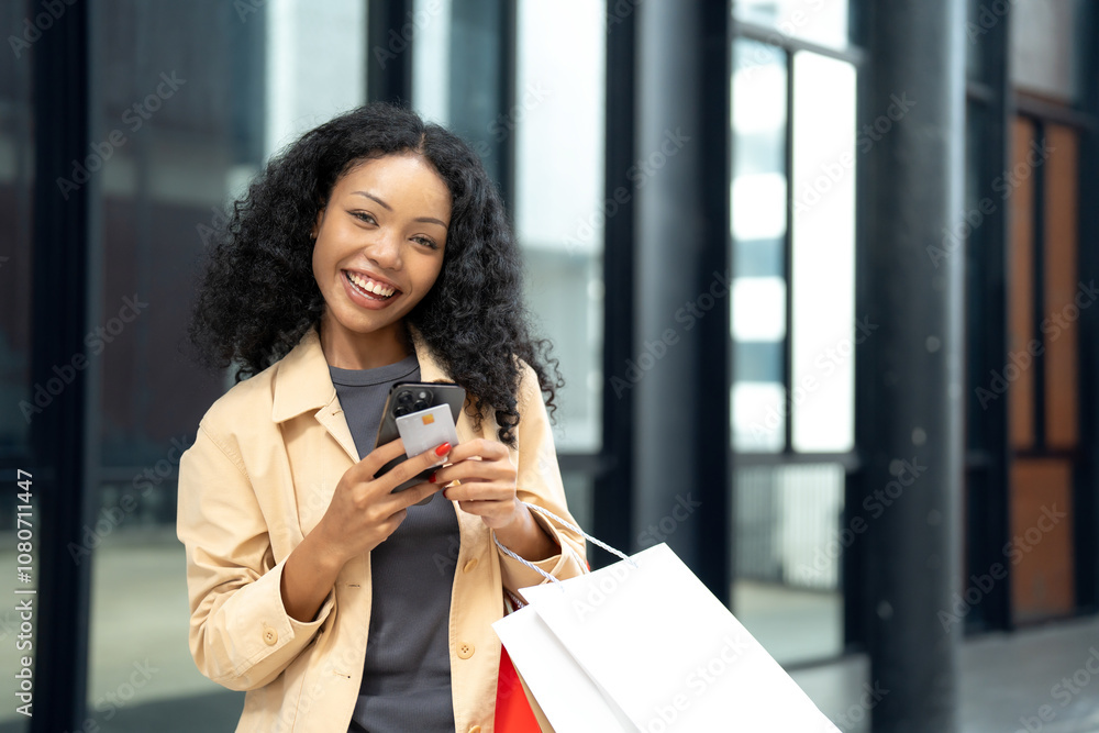 Fototapeta premium Happy woman holding shopping bags, a credit card, and a smartphone outdoors, representing online shopping and retail satisfaction.