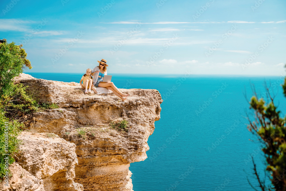 A woman and her child are sitting on a rock overlooking the ocean. The woman is using a laptop while the child is playing with a dog. A woman and her child are sitting on a rock overlooking the ocean. The woman is using a laptop while the child is playing with a dog.