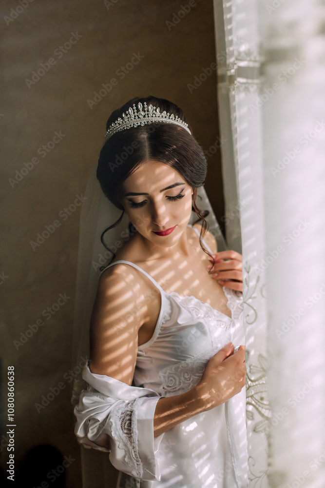 Fototapeta premium A woman in a white dress is standing in front of a window, with her arms crossed and a veil on her head. Concept of elegance and sophistication, as the woman is dressed in a wedding-like outfit