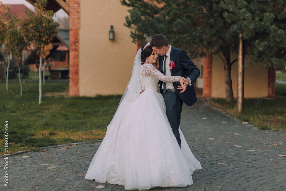 A bride and groom are dancing on a cobblestone path. The bride is wearing a white dress and the groom is wearing a blue suit