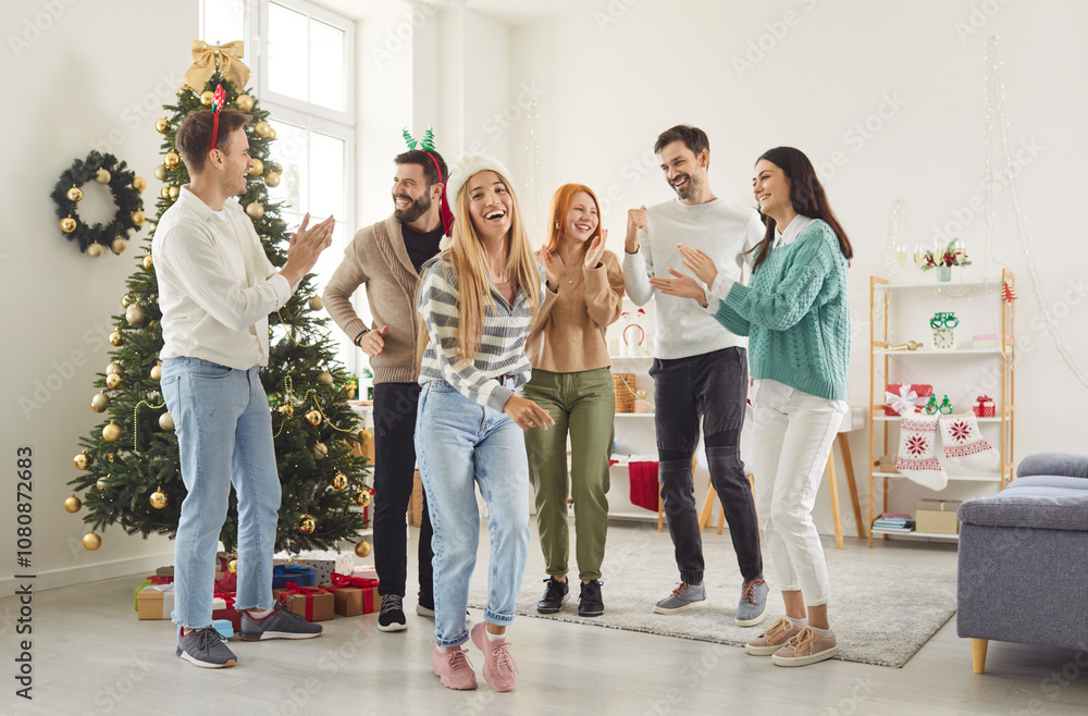 Fototapeta premium Happy smiling young people friends men and women having party, dancing in festive living room with decorated Christmas tree having fun celebrating New Year together. Winter holidays concept.