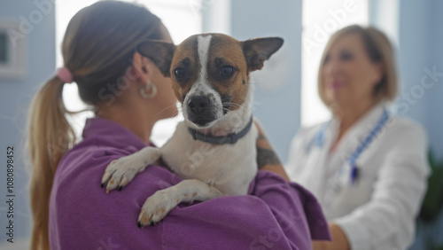 A middle-aged woman holds her dog in a veterinary clinic while another mature woman veterinary professional talks to them, showcasing a friendly indoor pet examination setting. photo