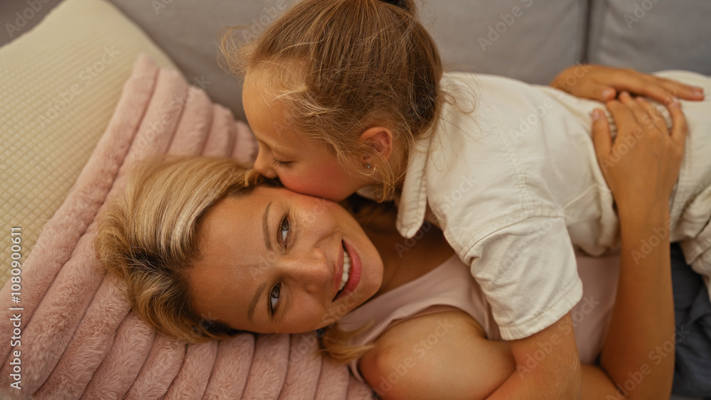 Blonde daughter gives a kiss to her mother while they lie together indoors on a cozy sofa in a loving and intimate moment Blonde daughter gives a kiss to her mother while they lie together indoors on a cozy sofa in a loving and intimate moment